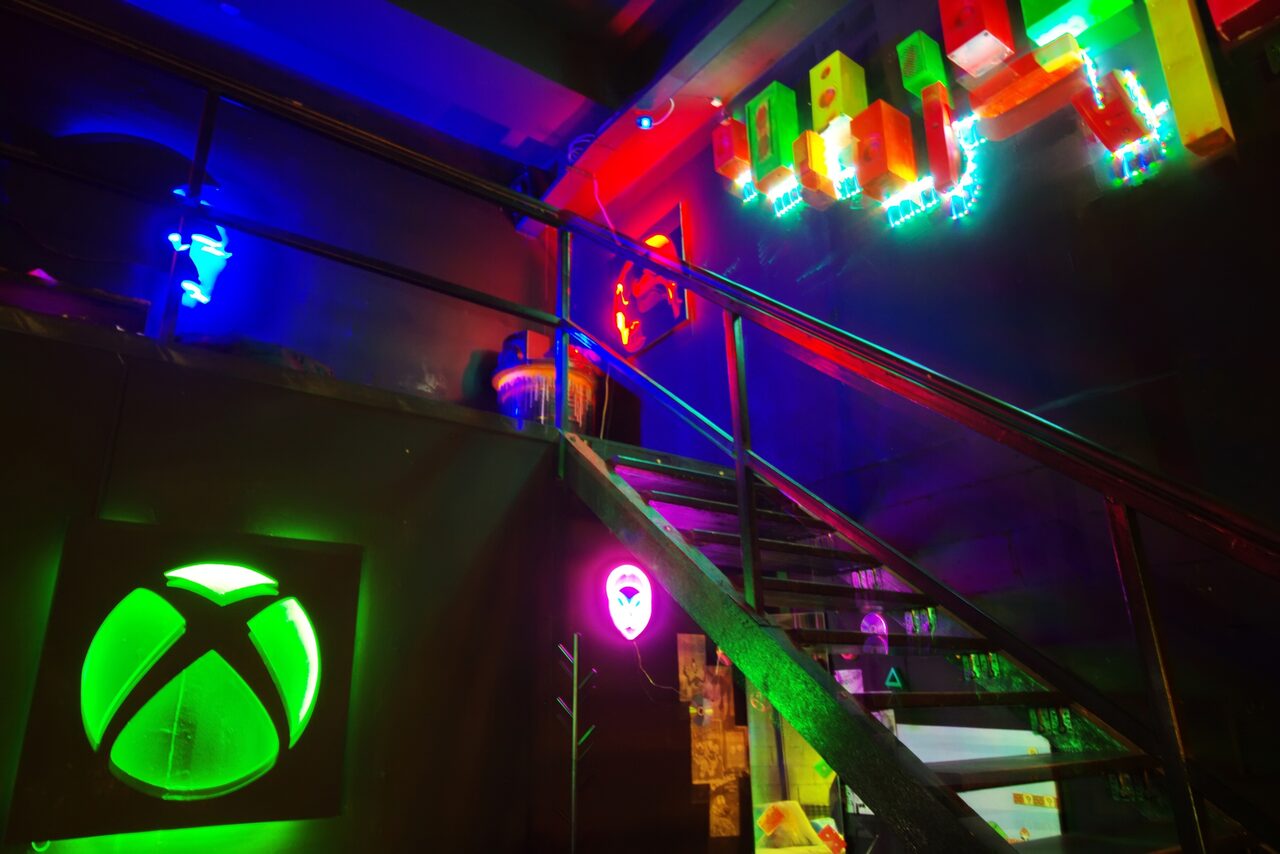 Арендовать Лофт Геймеры. Лестница на второй этаж, под ней неоновая подвеска с игровым логотипом и неоновая лампа. 