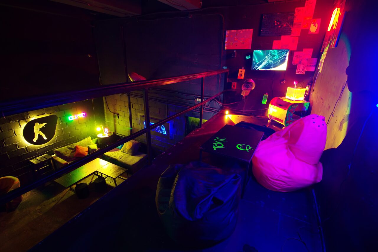 Арендовать Лофт Геймеры. Второй этаж лофта. К стене прикреплены небольшие картины с телевизором. На полу стоит небольшой столик с пуфами, в углу маленькая тумбочка с лампой. Также видно убранство первого этажа