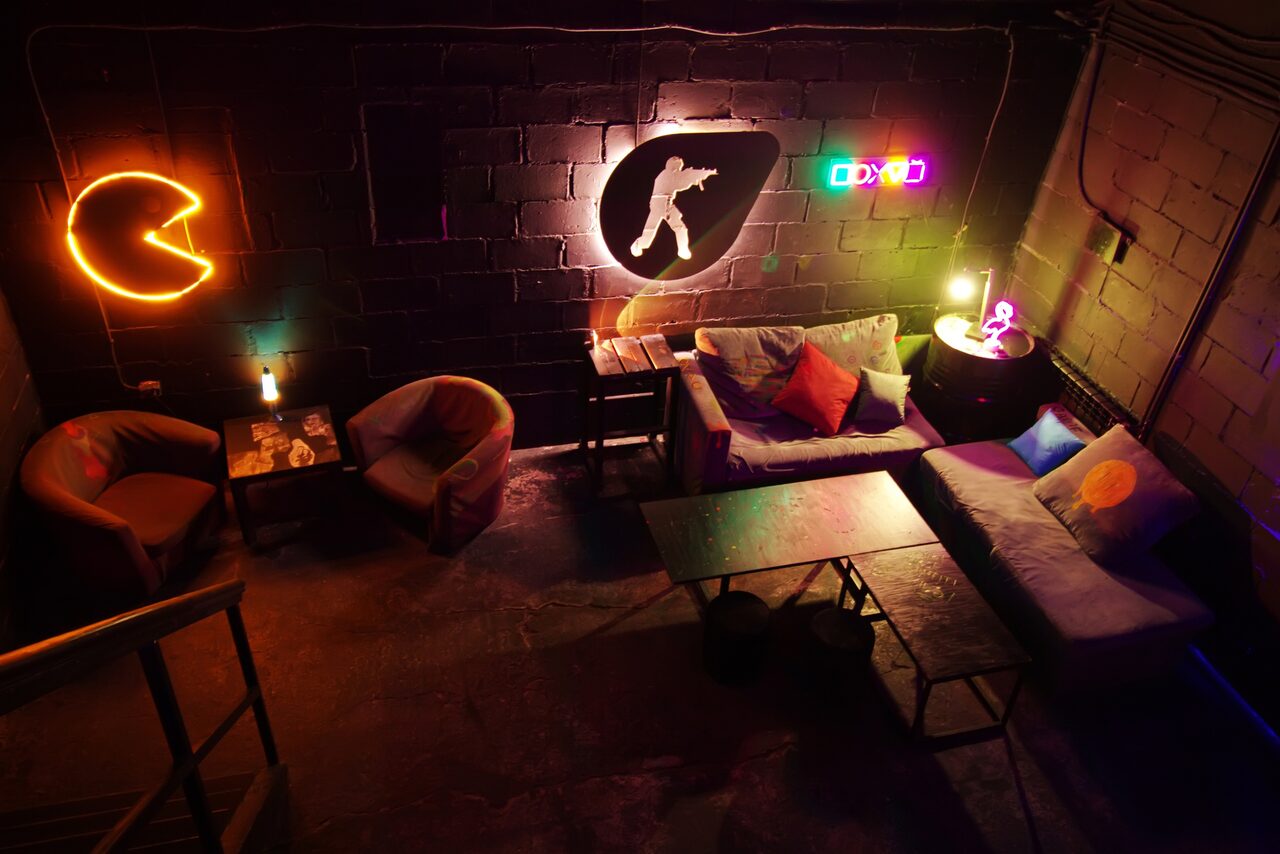 Арендовать Лофт Геймеры. Тёмный лофт. Неоновые подсветки, игровые логотипы на кирпичной стене. Несколько кресел, столов, диванчиков. Фотография сделана со второго этажа
