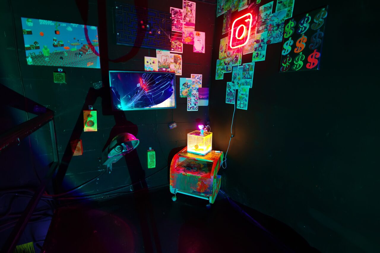 Арендовать Лофт Геймеры. Много различных изображений на стене и тематических иконок. Неоновая подсветка. Небольшая полка с лампой