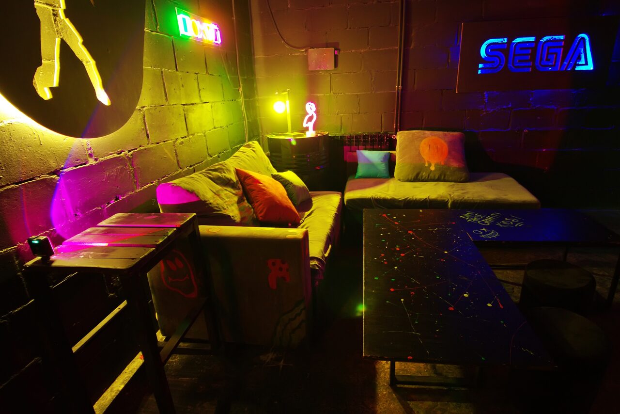 Арендовать Лофт Геймеры. Разрисованные диваны, стол. Бочка между диванами в углу. Неоновые изображения на стенах 