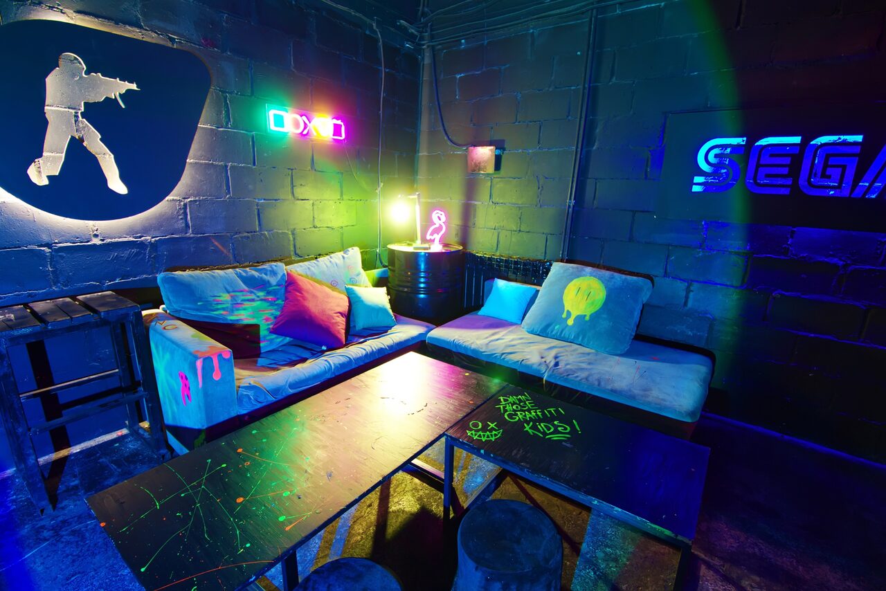 Арендовать Лофт Геймеры. Разрисованные диваны, стол. Бочка между диванами в углу. Неоновые изображения на стенах 