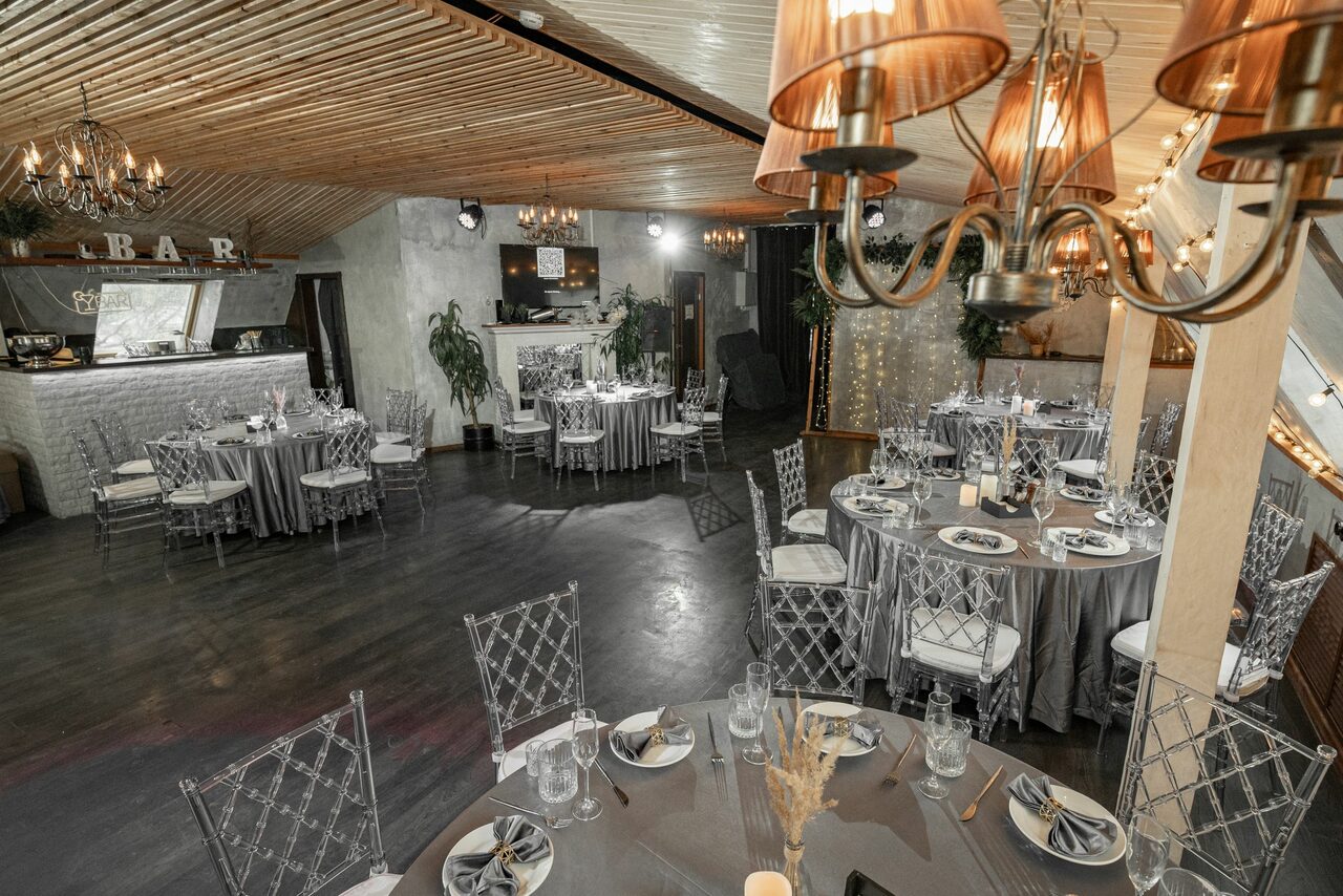Арендовать Loft - bar under closure in Peterhof. Свадебное пространство с круглой рассадкой, прозрачными стульями, бело-серебристой скатертью. Деревянный потолок, деревянный пол, декоративные растения. Зона бара с надписью «BAR». Люстры с лампами, подвесной свет.