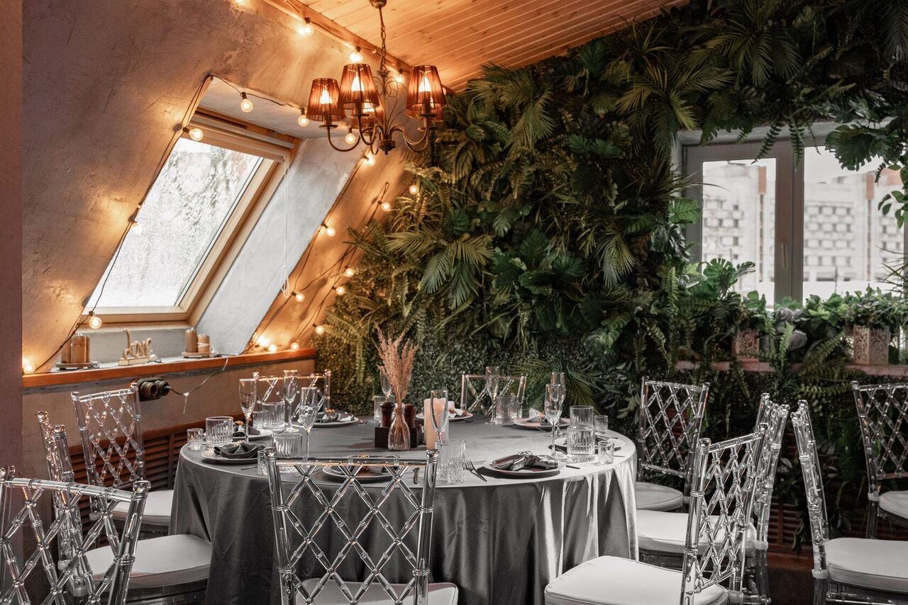 Арендовать Loft - bar under closure in Peterhof. Чердачное пространство с круглым столом, покрытым серой скатертью. Прозрачные стулья, накрытие на столе. На стене зелень. Потолочный светильник и гирлянда из лампочек. Косое окно с видом на улицу. Светлые деревянные панели на потолке.