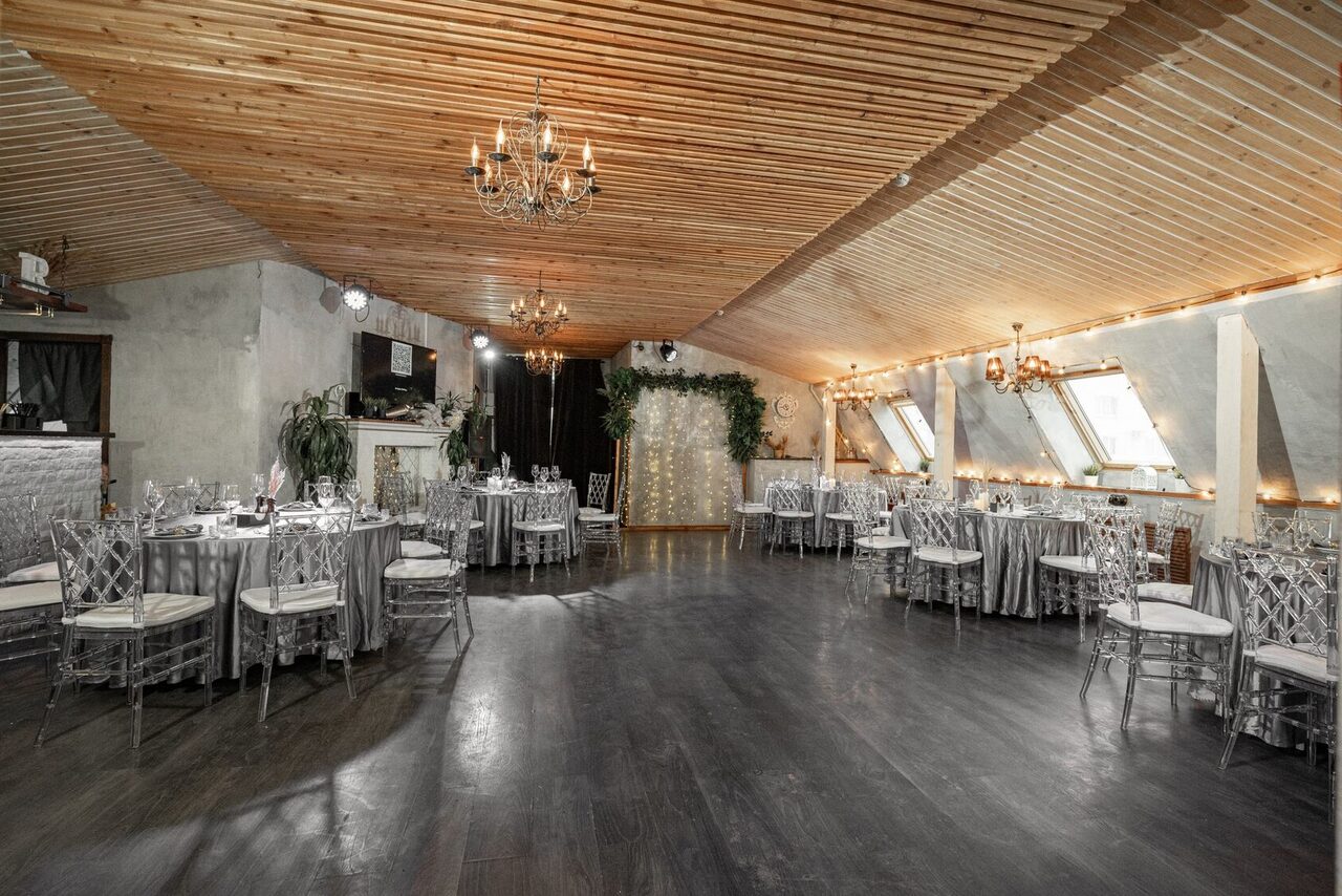 Арендовать Loft - bar under closure in Peterhof. Просторное помещение для мероприятий с деревянным потолком и люстрами, серыми стенами и тёмным полом. Обеденные столы с серыми скатертями и прозрачными стульями. У стены зелёные растения и световая гирлянда. Диагональные окна, мягкое освещение.