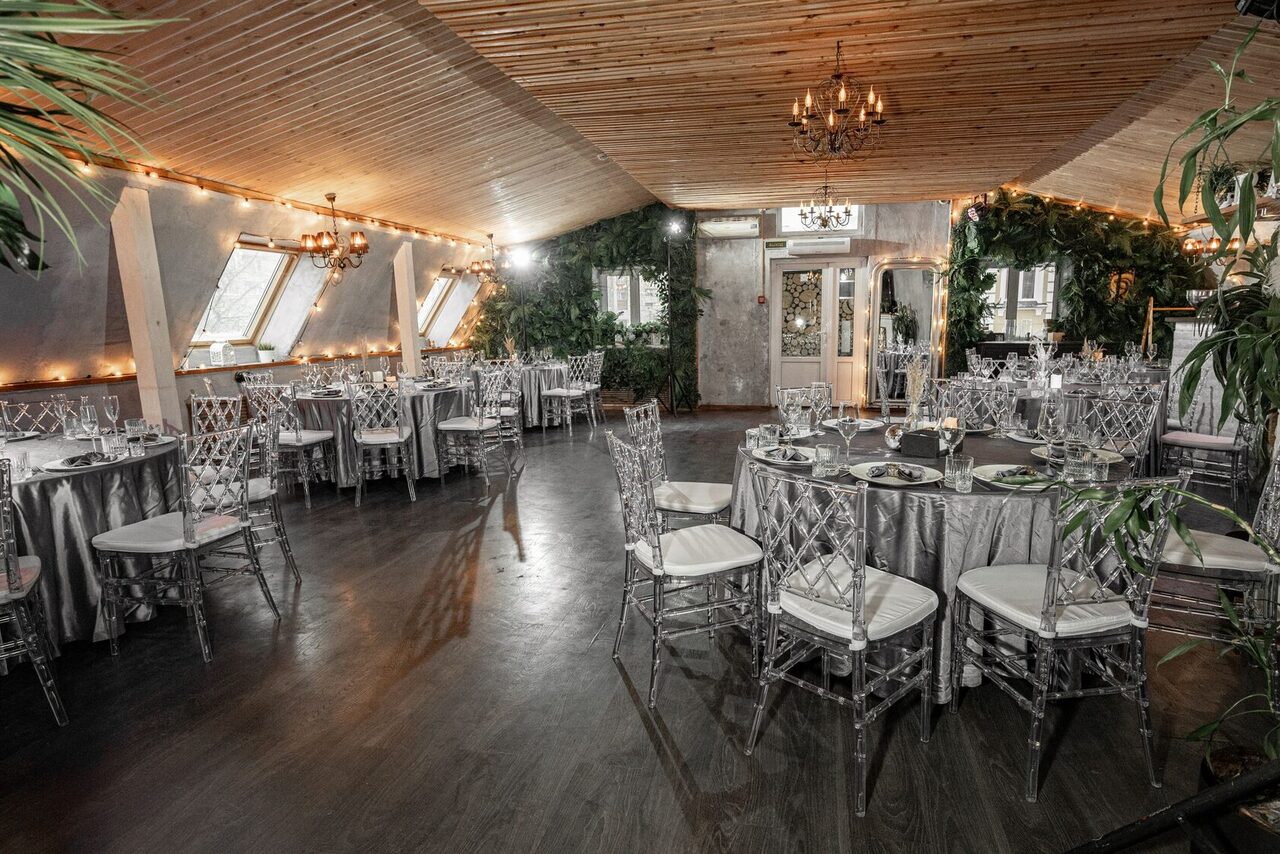 Арендовать Loft - bar under closure in Peterhof. Зал с деревянным потолком и тёмным полом, круглые столы с серыми скатертями и прозрачными стульями, оконные проёмы с наклоном, декор из зелени, люстры, подвесные светильники, гирлянды по периметру.