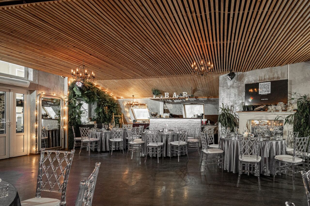 Арендовать Loft - bar under closure in Peterhof. Ресторан с деревянным решетчатым потолком и люстрами. Круглые столы с серебристыми скатертями и прозрачными стульями. Барная стойка на заднем плане. Зеркала, растения, светлая отделка стен. На стене вывеска с надписью "BAR".