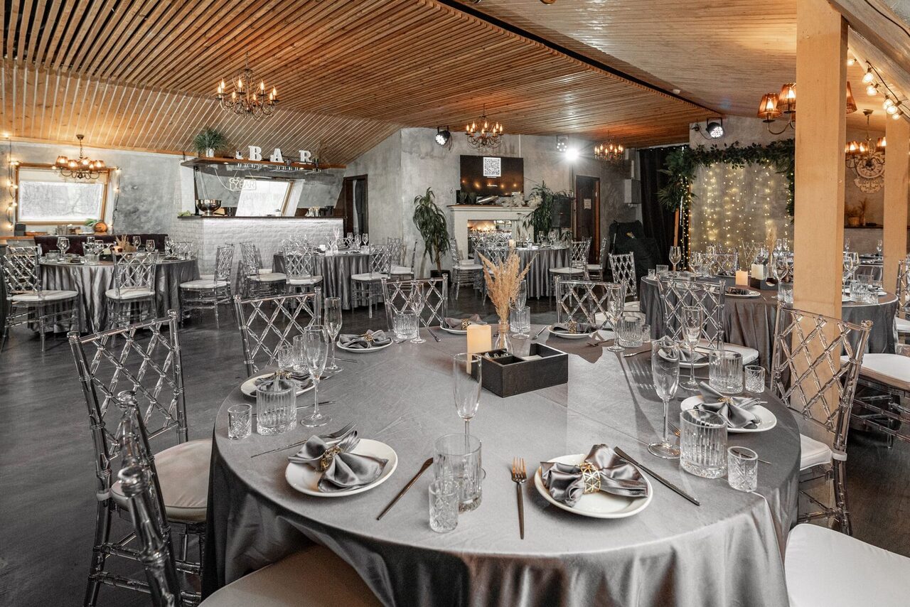 Арендовать Loft - bar under closure in Peterhof. Просторное мероприятие с круглым столами, сервированными столовыми приборами и стеклянной посудой. Стулья прозрачные, потолок из деревянных панелей. Освещение теплое, люстры. Стены серые, бар с надписью "BAR". Декор из растений и световых гирлянд.