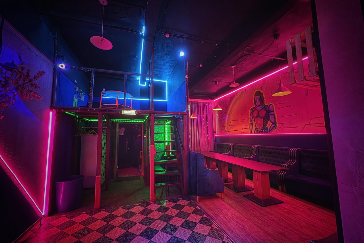 Арендовать Лофт в стиле Диско. Клубное пространство с неоновым освещением; на стенах росписи героя. Пространство с лестницей на второй ярус, зона отдыха с длинным столом и мягкими диванами. Черно-белая клетчатая плитка на полу и деревянные покрытия. Светильники на потолке.