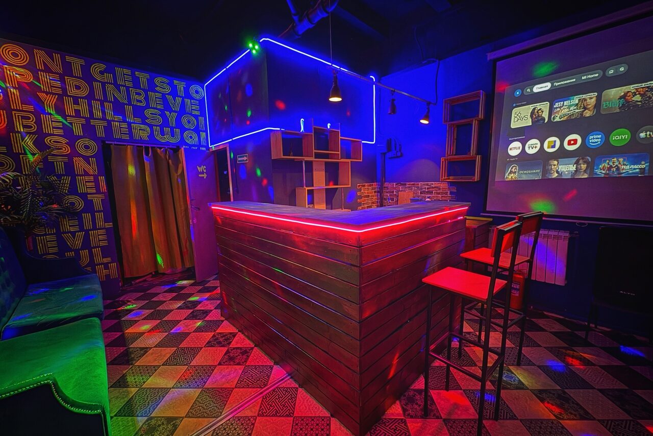 Арендовать Лофт в стиле Диско. Барная зона с деревянной стойкой, освещенной красными и синими неоновыми лампами. В помещении высокие столы и стулья, проектор с изображением экранного интерфейса. На полу разноцветная плитка. На стене неоновая надпись.