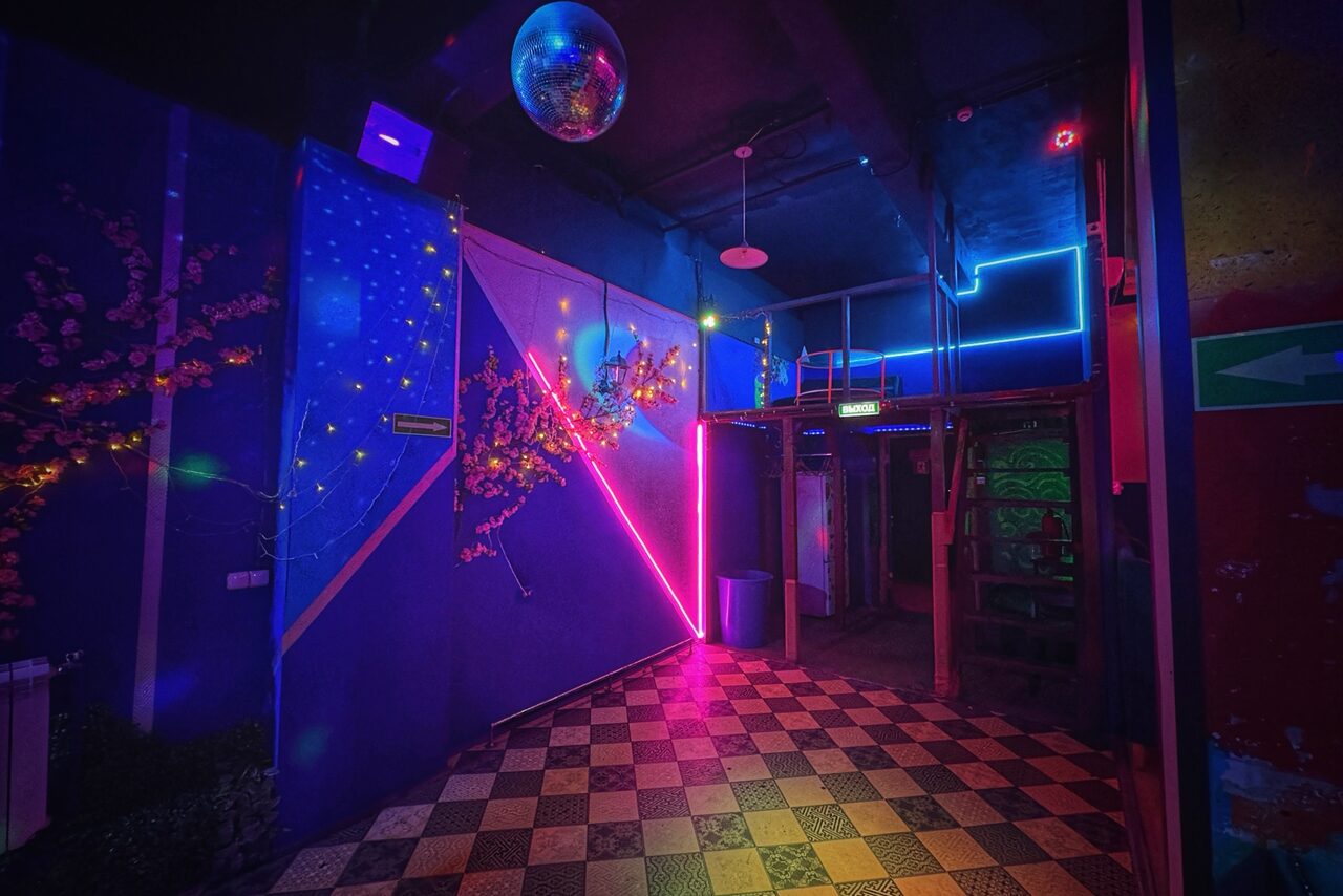 Арендовать Лофт в стиле Диско. Темное помещение с неоновым освещением; геометрическая планировка; диско-шар на потолке; декоративные элементы с розовыми и синими светодиодами; клетчатый пол; неоновая стрелка-указатель на стене; две входные двери; металлическая лестница.