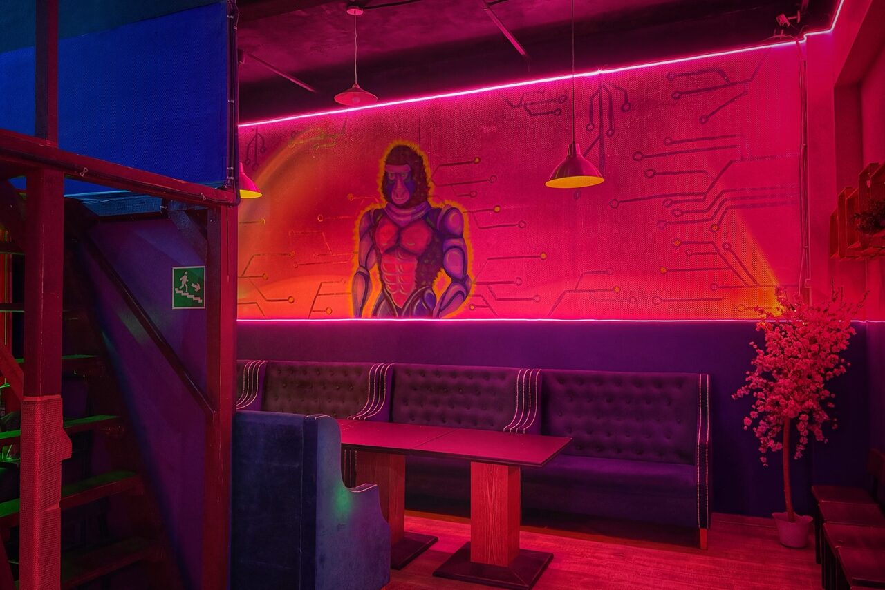 Арендовать Лофт в стиле Диско. Интерьерная зона с неоновым освещением, настенными рисунками, включая фигуру человека в стиле киберпанк. Мягкие диваны, столы из светлого дерева, подвесные светильники. Стены украшены рисунками с имитацией электронных схем.