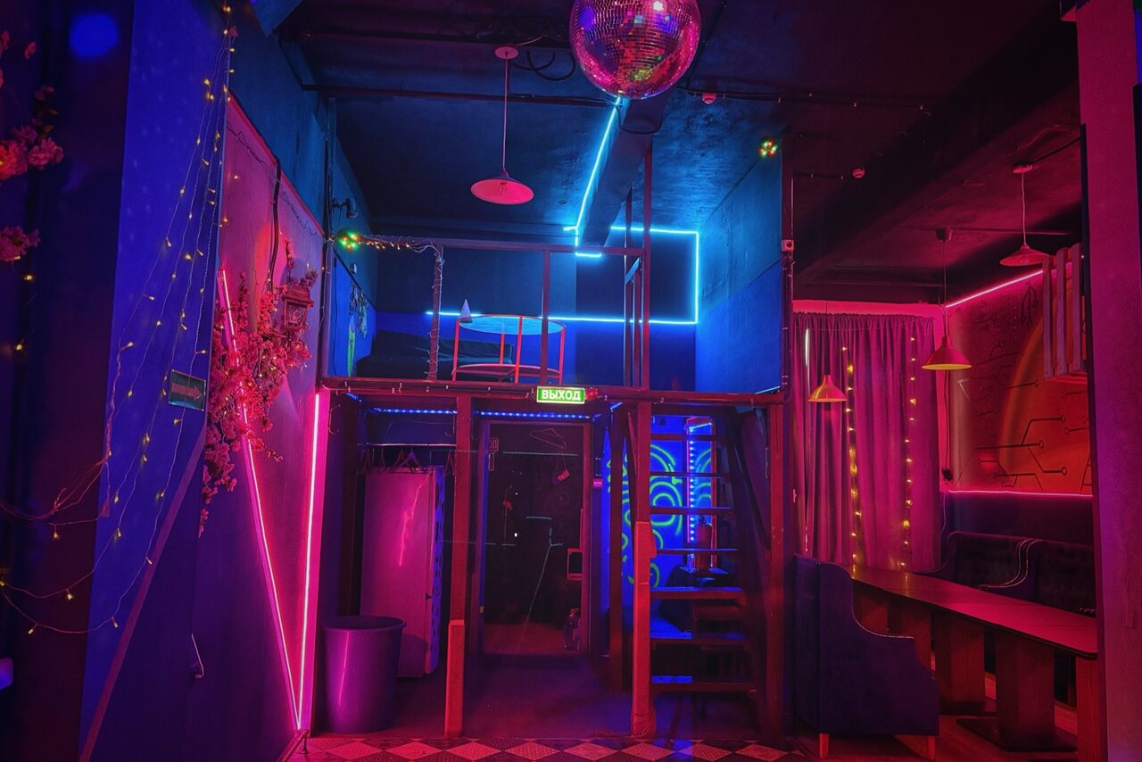 Арендовать Лофт в стиле Диско. Ночное пространство с неоновой подсветкой, многоуровневой планировкой и металлической лестницей. Стены окрашены в синий, розовый и фиолетовый. Есть мягкие диваны, шторы и диско-шар. На стене растительные декорации и светодиодные ленты.