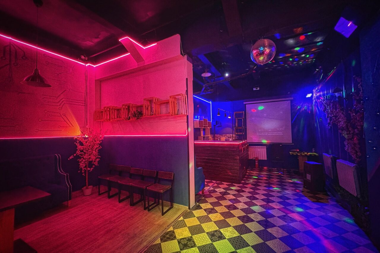 Арендовать Лофт в стиле Диско. Лофт-пространство с барной стойкой, деревянными стульями и скамейками. Черно-клетчатый пол, проекционный экран. Красное неоновое освещение, диско-шар на потолке. Декоративные растения. Стены окрашены в темные и розовые тона.