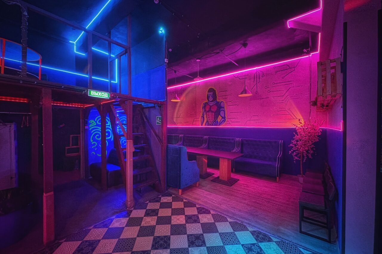 Арендовать Лофт в стиле Диско. Лофт-пространство с яркой неоновой подсветкой, лестницей на второй уровень, фреской на стене. Мебель: большие диваны и столы. Пол: шахматная плитка и паркет. Освещение разноцветное: розовое, синее, фиолетовое. Видны двери с надписями "Вход".