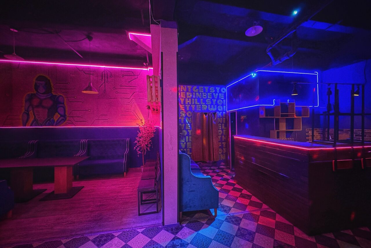 Арендовать Лофт в стиле Диско. Клубное пространство с неоновым освещением в фиолетовых и синих тонах. Зона барной стойки; на стенах рисунки и надписи. В углу диван и стол, рядом высокие стулья. Пол с узорчатым покрытием; присутствуют декоративные элементы.