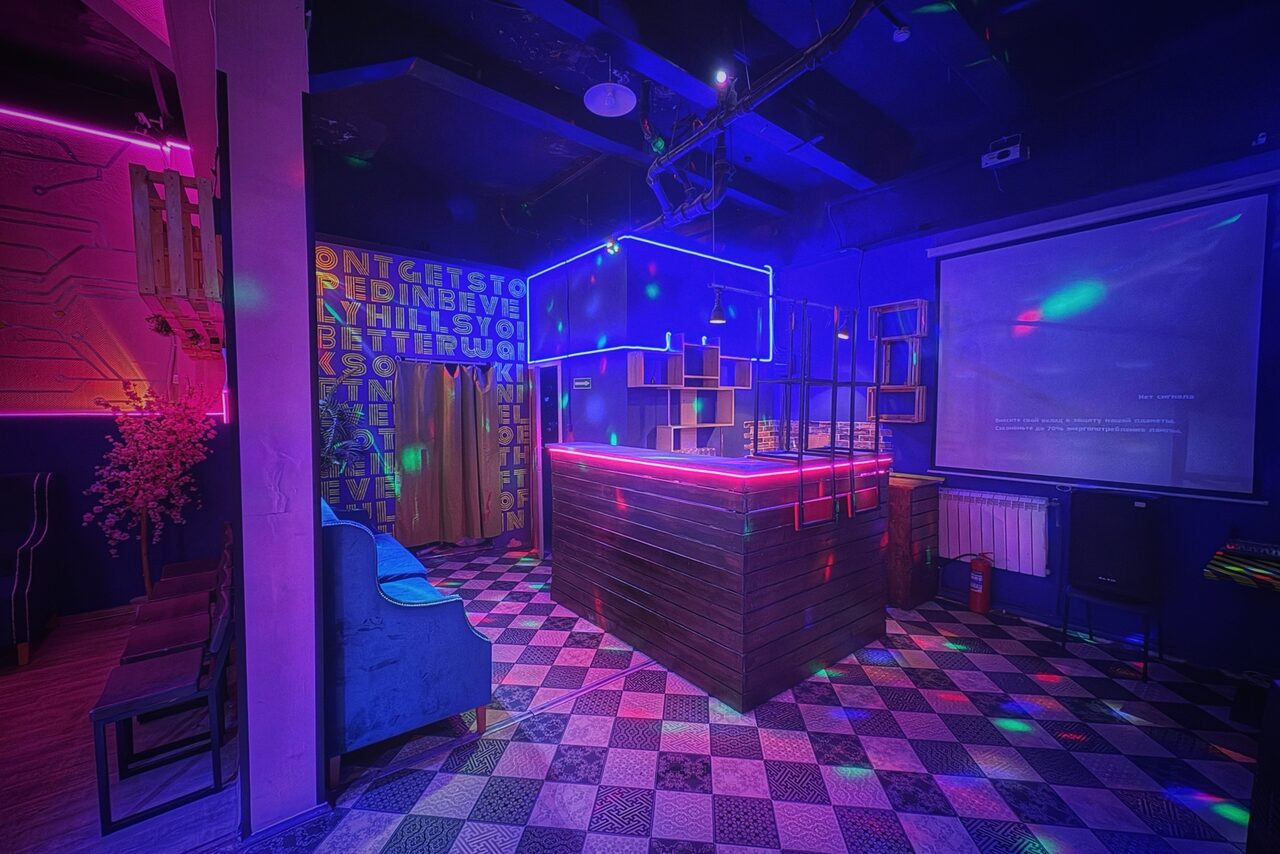 Арендовать Лофт в стиле Диско. Клубное пространство с барной стойкой, окруженной стальными рейками. Пол плиточный, узорчатый, черно-белый. Стены темные с неоновыми надписями. Имеется проектор и экран. Освещение разноцветное, неоновое. Мебель: кресла и столики.