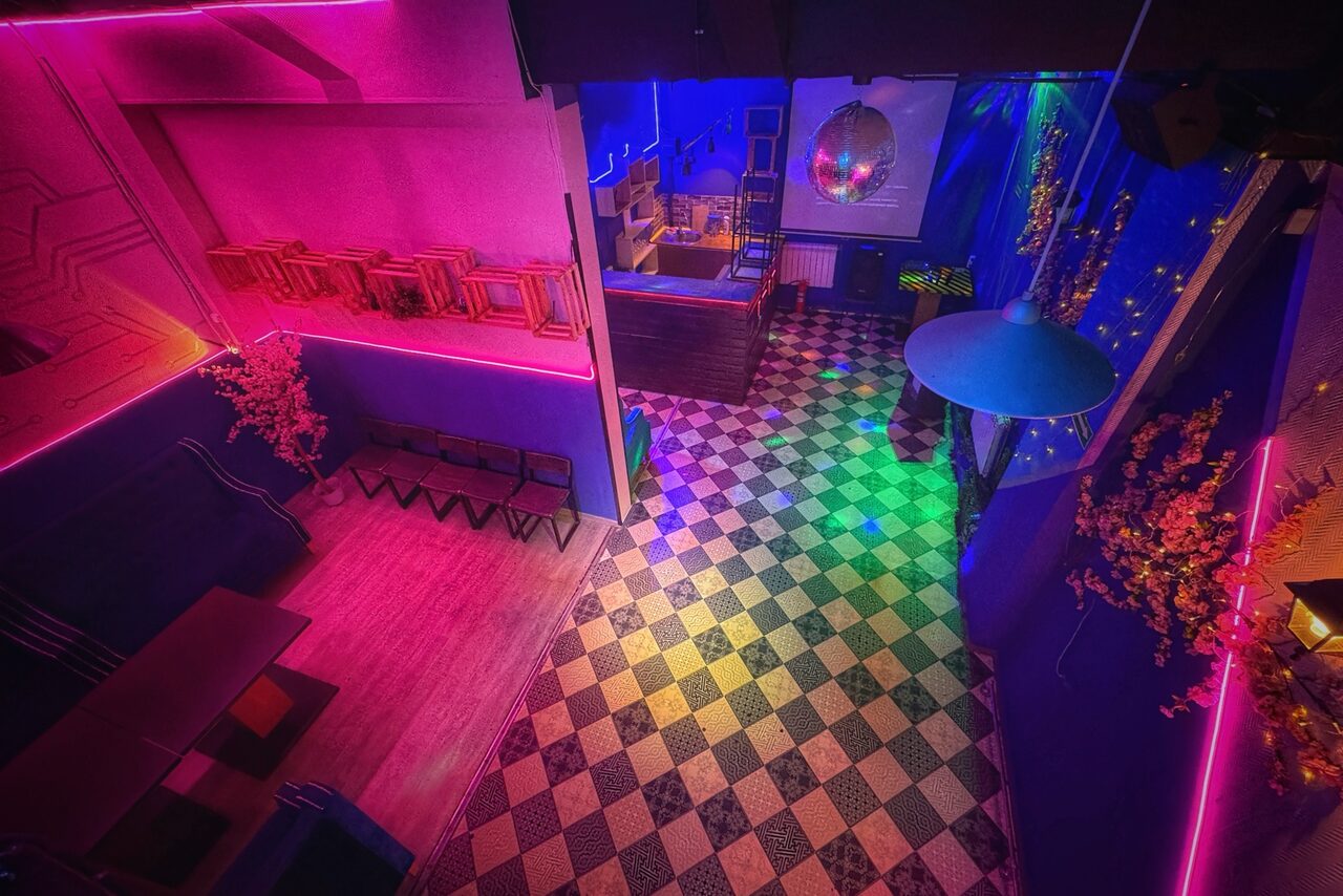 Арендовать Лофт в стиле Диско. Пространство с высокими потолками; мозаичный пол с шахматным узором; стены с розовой и синей подсветкой; столы и лавки вдоль стен; барная стойка; диско-шар; декоративные элементы на стенах.