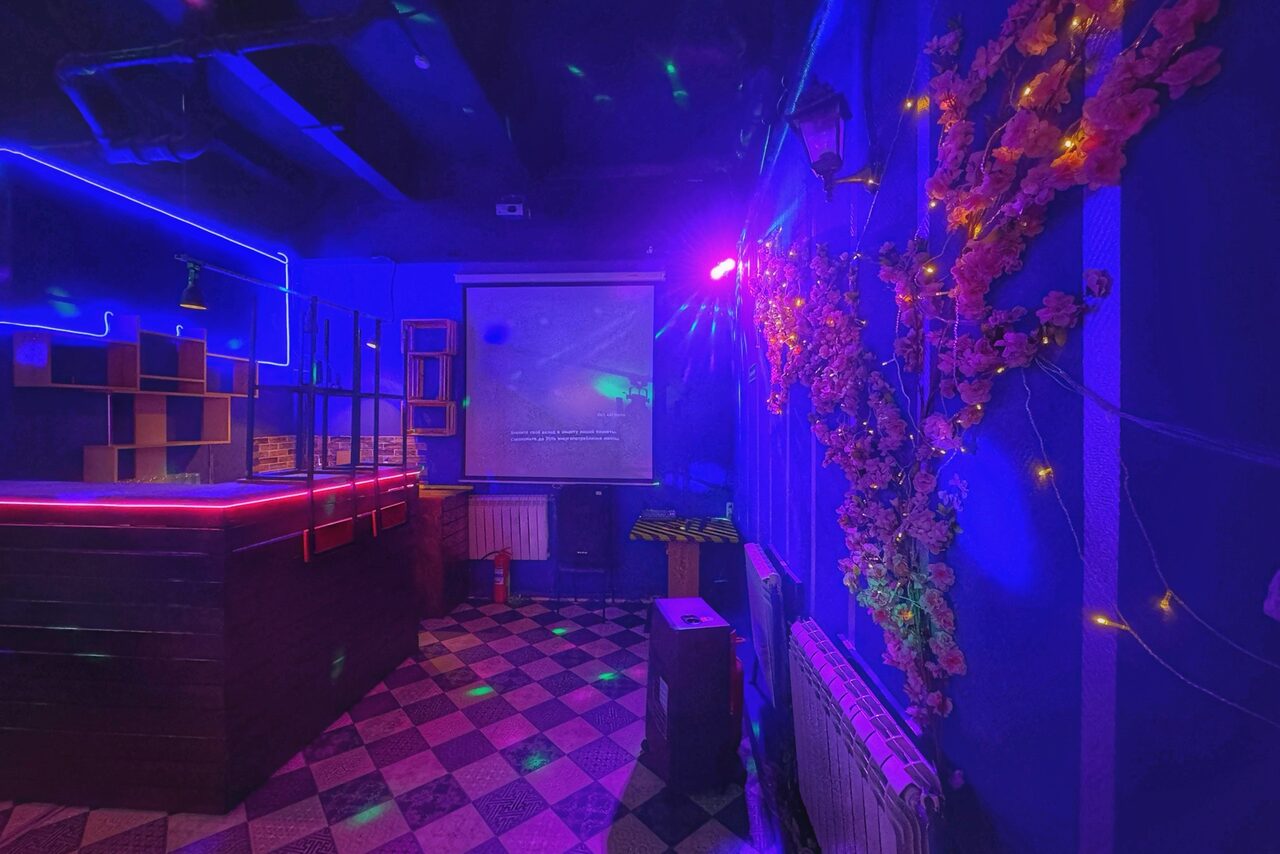 Арендовать Лофт в стиле Диско. Караоке-зал с барной стойкой и проектором, цветное освещение, черно-белая клетчатая плитка на полу, стены украшены гирляндами и цветами, деревянные элементы мебели, на стене экран с текстом.