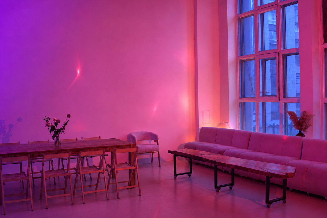 Арендовать  Light loft with light music and a bar. Просторное помещение с высоким потолком, большой диван вдоль стены, деревянный стол с несколькими стульями, декоративные растения, крупные окна, розово-фиолетовая подсветка, минималистичный современный стиль.