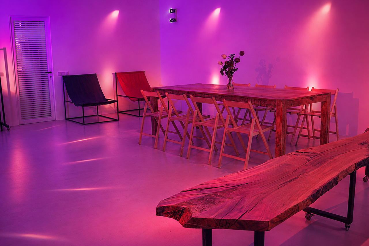Арендовать  Light loft with light music and a bar. Просторная комната с бетонным полом и фиолетовым освещением. Деревянный стол с букетом и складными стульями, в углу кресла на каркасе. На стене лампы и двери с жалюзи. Декоративная деревянная скамья.