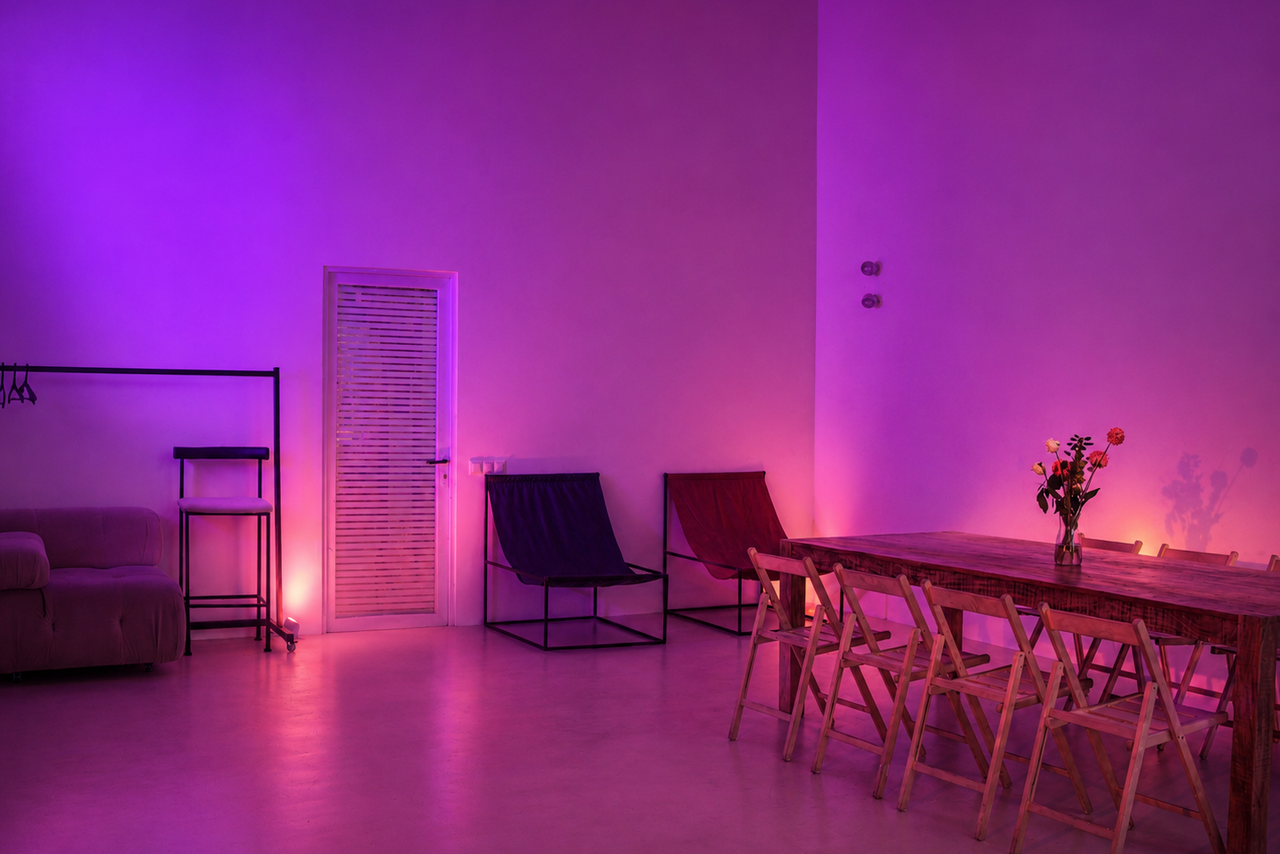 Арендовать  Light loft with light music and a bar. Просторная комната с фиолетовой и розовой подсветкой. Деревянный стол с цветами и деревянными складными стульями. Диван, стеллаж и шезлонги справа. Дверь с жалюзи. Пастельные тона, гладкие поверхности.