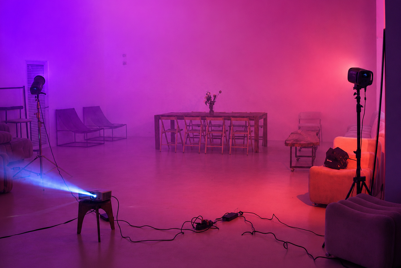 Арендовать  Light loft with light music and a bar. Просторное помещение с бетонным полом и фиолетово-розовым освещением. В центре деревянный стол с цветами и стульями, вокруг диваны и кресла. Слева софиты, справа проектор с синим светом. Пауки кабелей на полу.