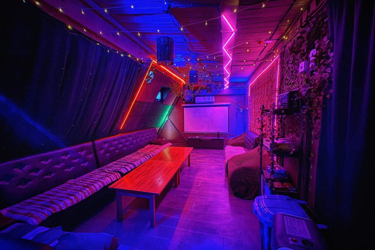 Арендовать Лофт-бар. Караоке-комната с мягкими диванами и пуфами, длинный деревянный стол, на стене проекционный экран. Неоновая подсветка пурпурного и синего цвета, гирлянды. Темные занавески, стены декорированы под камень. Пол плиточный.
