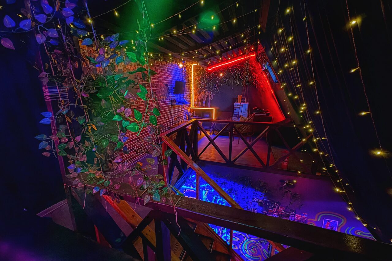 Арендовать Лофт-бар. Лофт-пространство с кирпичными стенами, деревянными перилами и балконом. Потолок украшен гирляндами. Зона освещена разноцветными неоновыми лампами. На мебельной полке несколько предметов, на стене плоский экран. Растения свисают с перил.