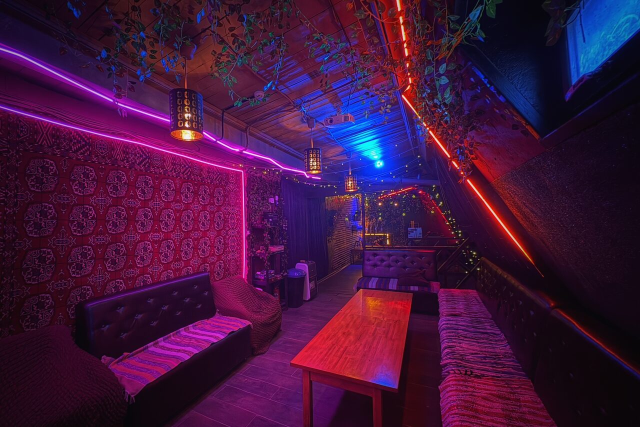Арендовать Лофт-бар. Лофт с мягкой мебелью, включая диваны и кресла, деревянный стол. Стены с узорчатым ковром, освещение неоновыми и гирляндными огнями, разноцветные светильники на потолке. Потолочные балки декорированы растениями. Темные тона, яркие цветные акценты.