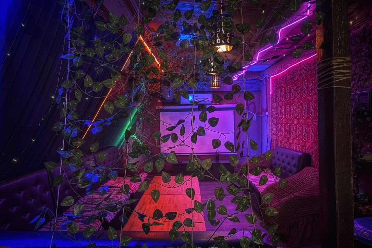 Арендовать Лофт-бар. Камерное пространство с мягкими диванами по периметру, деревянным столом в центре. Потолочный светильник и неоновая подсветка различных цветов. На переднем плане декоративные растения. В глубине стена с узорчатым обоями, проекционный экран.