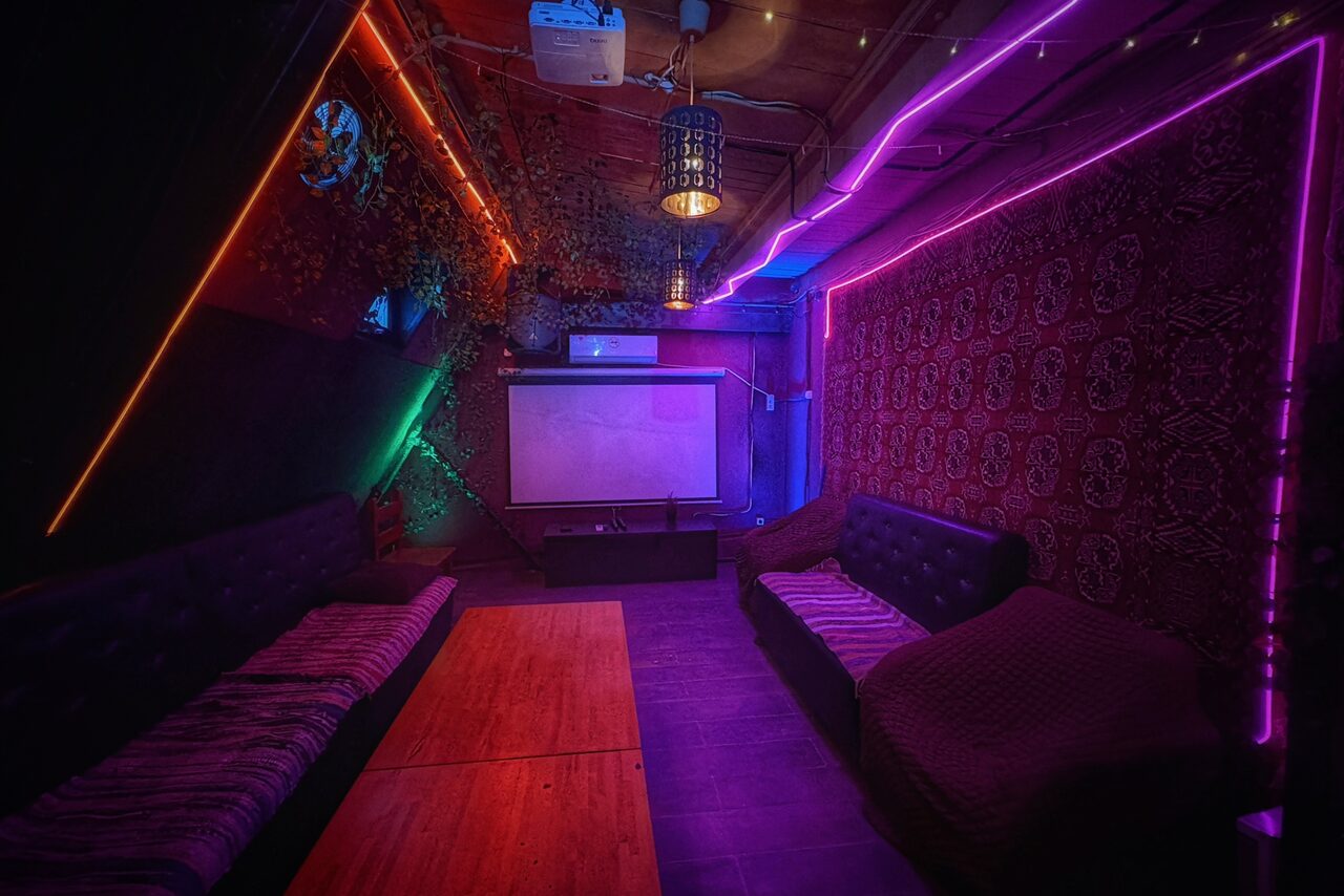 Арендовать Лофт-бар. Треугольное помещение с темными стенами и потолком, обрамленное яркой неоновой подсветкой. Два черных дивана, деревянный стол, проектор и экран. Разноцветные декоративные элементы. Теплые тона с элементами фиолетового и красного цветов.