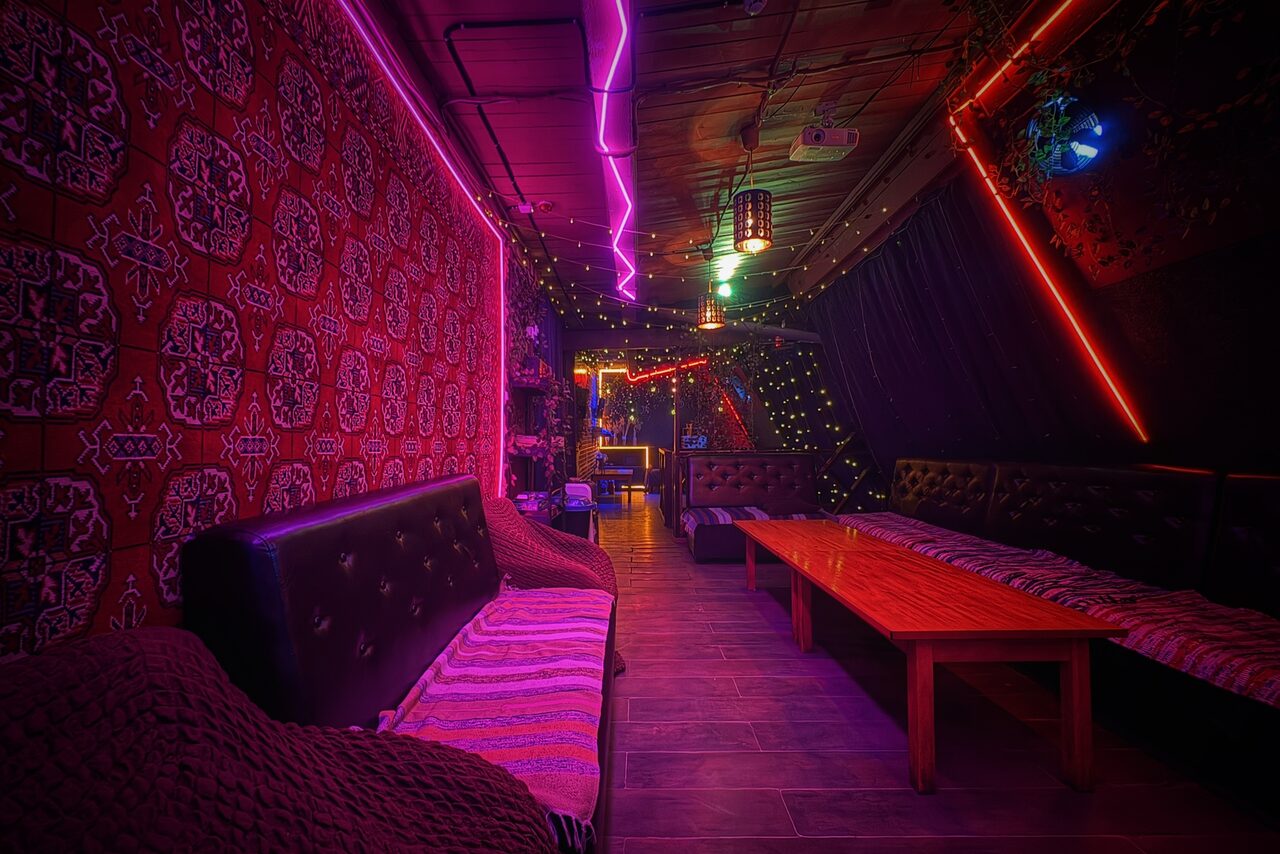 Арендовать Лофт-бар. Узкое помещение с мягкими диванами и столом вдоль стены. Разноцветное неоновое освещение, деревянный потолок, плиточный пол. Стены украшены узорчатыми тканями и гирляндами. Тёмные оттенки мебели. Подушки и пледы на диванах.