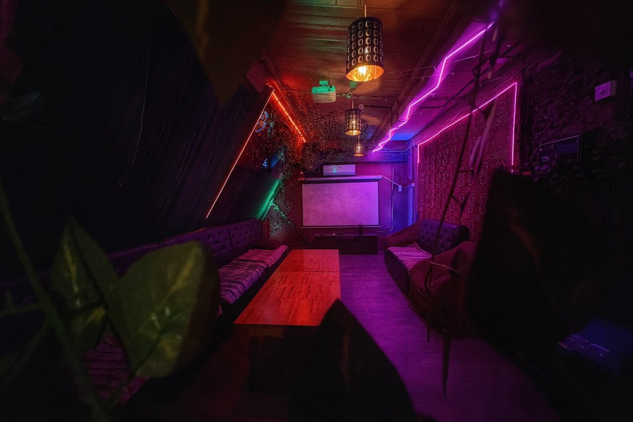 Арендовать Лофт-бар. Лофт пространство с низким освещением; на стенах светодиодные ленты разных цветов; потолочные подвесные светильники; мягкие диваны вдоль стен; на полу темное покрытие; в центре низкие столы; на стене экран; растительность на переднем плане.