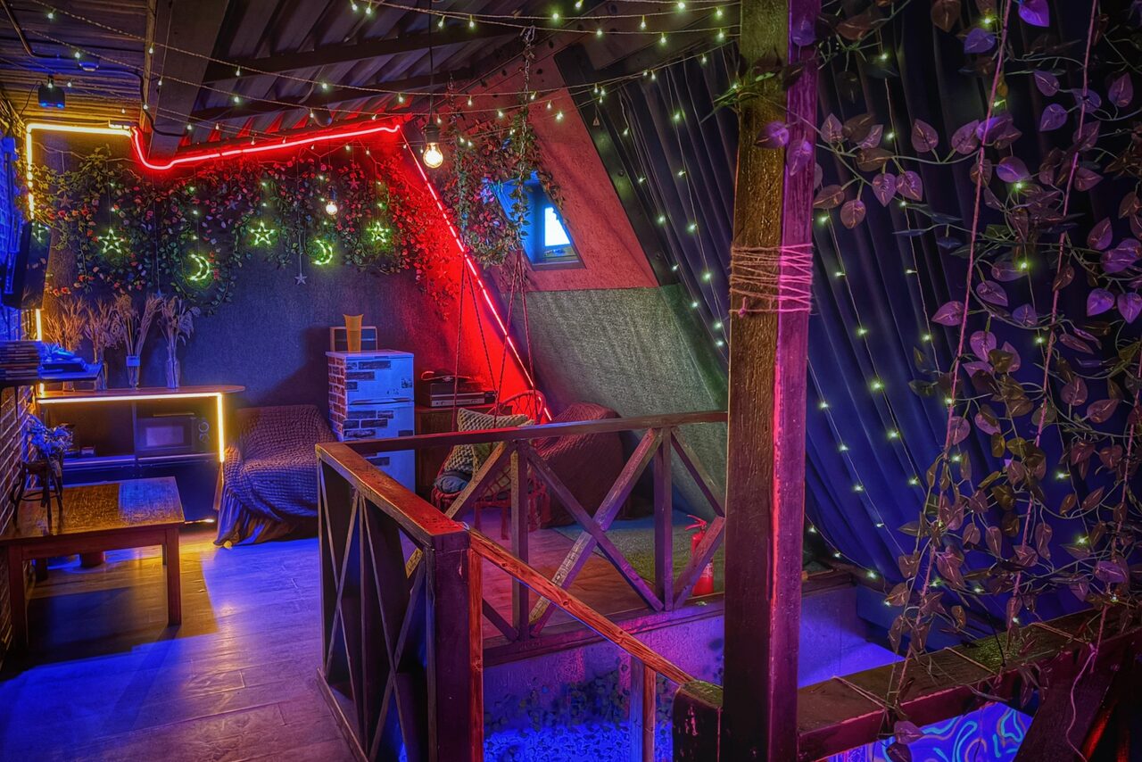 Арендовать Лофт-бар. Чердак с деревянными элементами и балюстрадой, отделка в стиле лофт. Диван с пледом, полки с предметами, телевизор. Яркое неоновое освещение, гирлянды и декоративные звезды. Занавески с растительным оформлением. В помещении царит разноцветное освещение.
