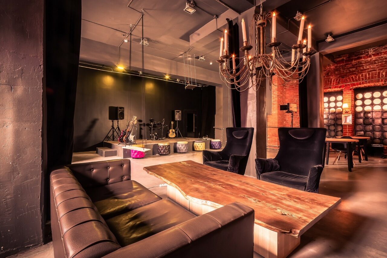 Арендовать Music loft (Мьюзик лофт). Лофт пространство со сценой, светильники и звукосниматели, чёрные стены и кирпич, кожаный диван, деревянный стол, два черных кресла, большая люстра, свет в виде свечей, винтовая лестница в углу, текстильные подушки на подиуме на сцене.