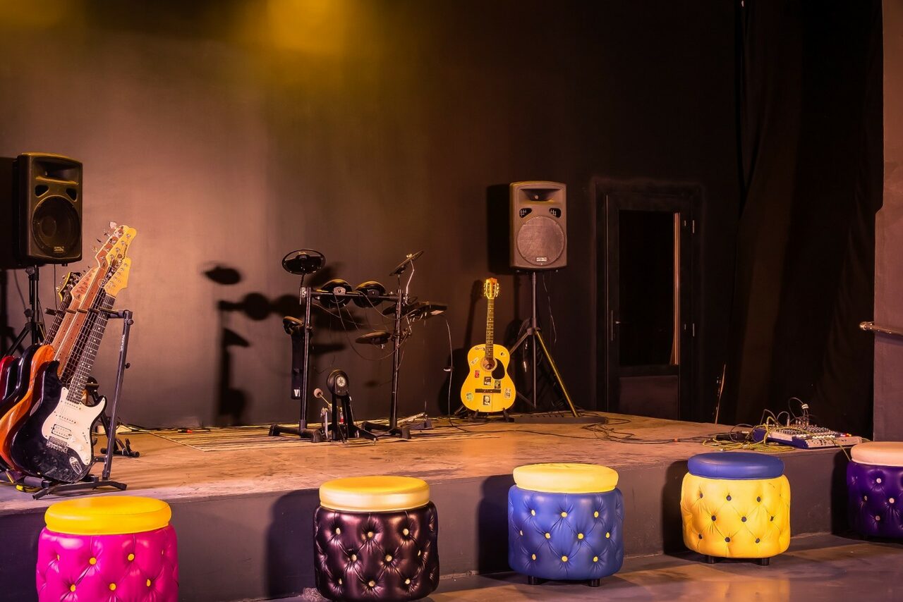 Арендовать Music loft (Мьюзик лофт). Сцена с черными стенами, на которой расположены музыкальные инструменты: две электрогитары, электронная ударная установка, колонка. Перед сценой разноцветные круглые пуфы. Освещение теплое, мягкое.