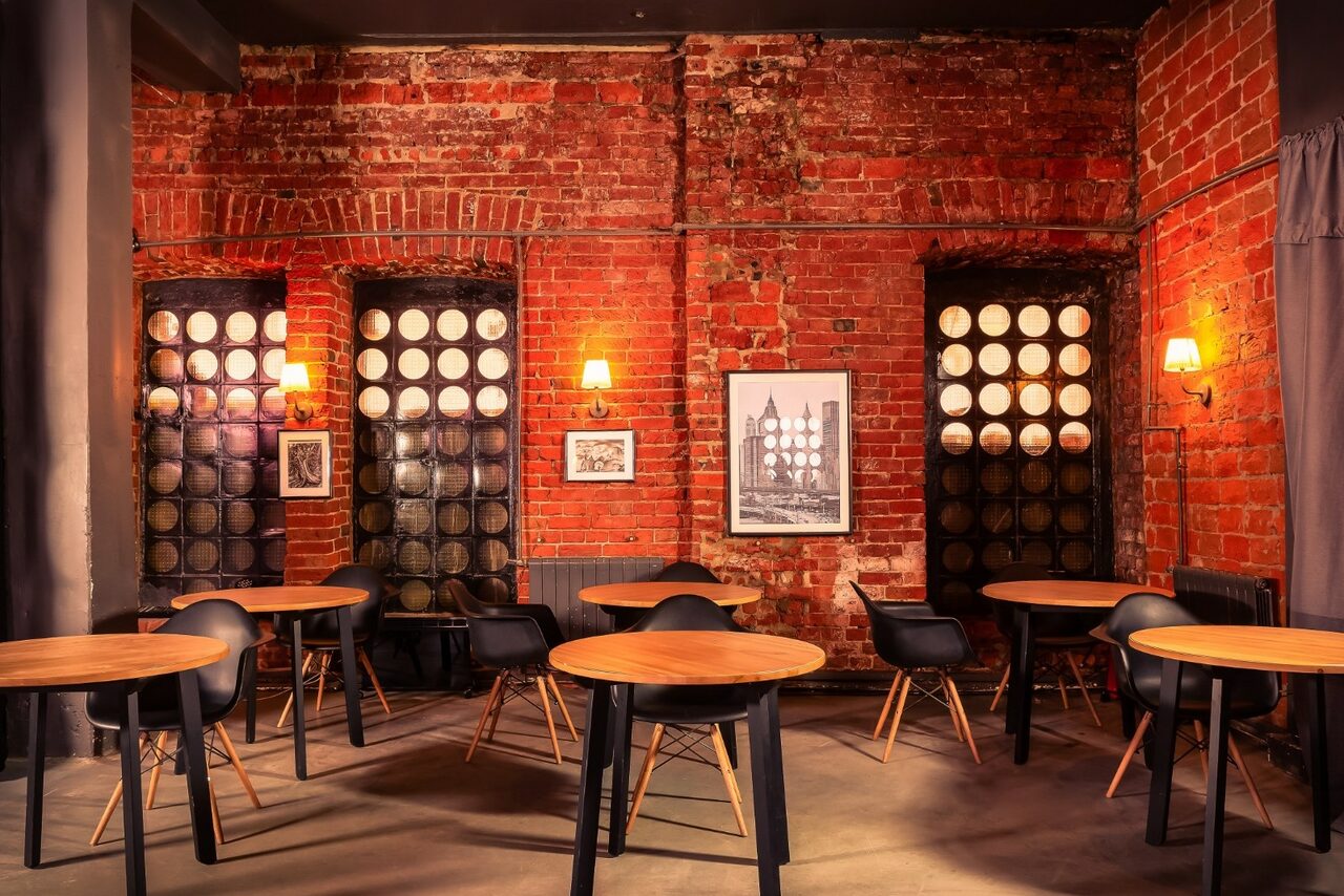 Арендовать Music loft (Мьюзик лофт). Интерьер с кирпичными стенами, круглые деревянные столы с черными стульями, панорамные окна с круглым остеклением. На стенах висят картины и светильники. Пол бетонный, свет теплый.