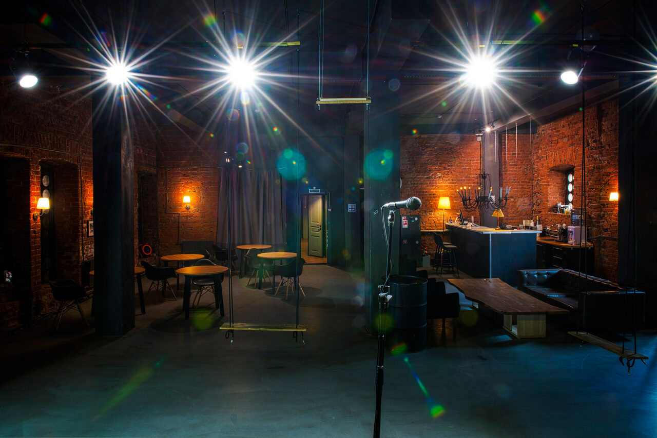 Арендовать Music loft (Мьюзик лофт). Лофт-пространство с кирпичными стенами, высокими потолками и промышленным освещением. В центре стойка с микрофоном. Есть диваны, круглые столы и барная стойка. Освещение яркое, зонирующее. Потолок из металлических конструкций. Цвета темные с акцентами.