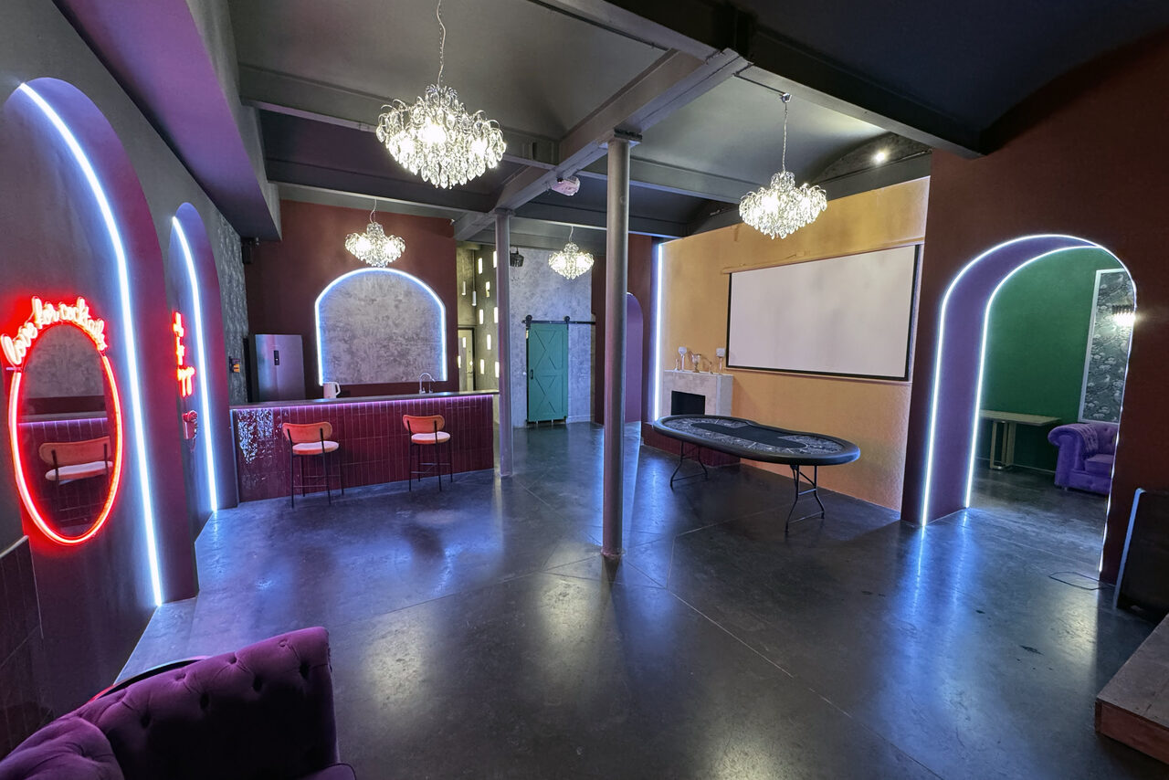 Арендовать Белый лофт с яркой мебелью. Просторное помещение с черным потолком и полом, украшенное сверкающими люстрами. В центре расположен длинный стол. Справа - белый экран для проектора. Ниша с барной стойкой и стульями. На стенах неоновые вывески, круглое зеркало с подсветкой.