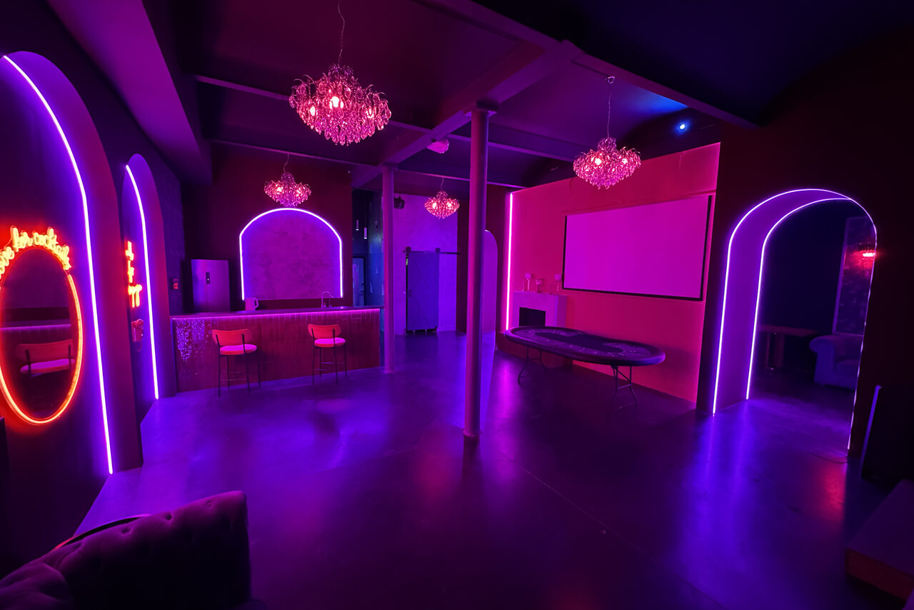 Арендовать Белый лофт с яркой мебелью. Тёмное помещение с неоновым освещением фиолетового и розового цветов. Барная стойка с двумя стульями, круглый стол, подвесные светильники. На стене белый прямоугольник, напоминающий экран. Арочные проёмы и бетонный пол.