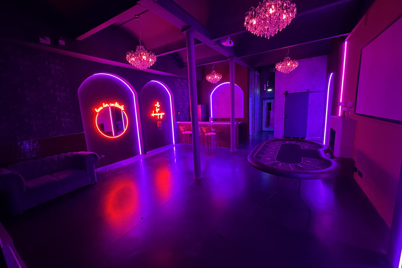 Арендовать Белый лофт с яркой мебелью. Современное пространство с неоновым освещением, тремя арками на стене, диваном слева, барной стойкой и табуретами посередине, покерным столом справа, люстрами на потолке. На стенах неоновые знаки. Цвета: фиолетовый, розовый.