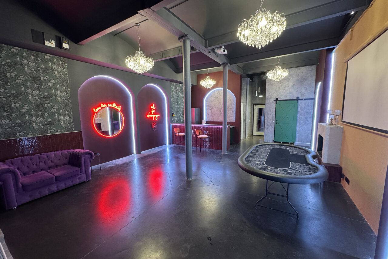 Арендовать Белый лофт с яркой мебелью. Просторная комната с высоким потолком, стол для игры, фиолетовый диван, барная стойка с табуретами, неоновая подсветка, декоративные арки, цветочные обои, зелёная раздвижная дверь, большие люстры. Пол тёмный, стены разноцветные.