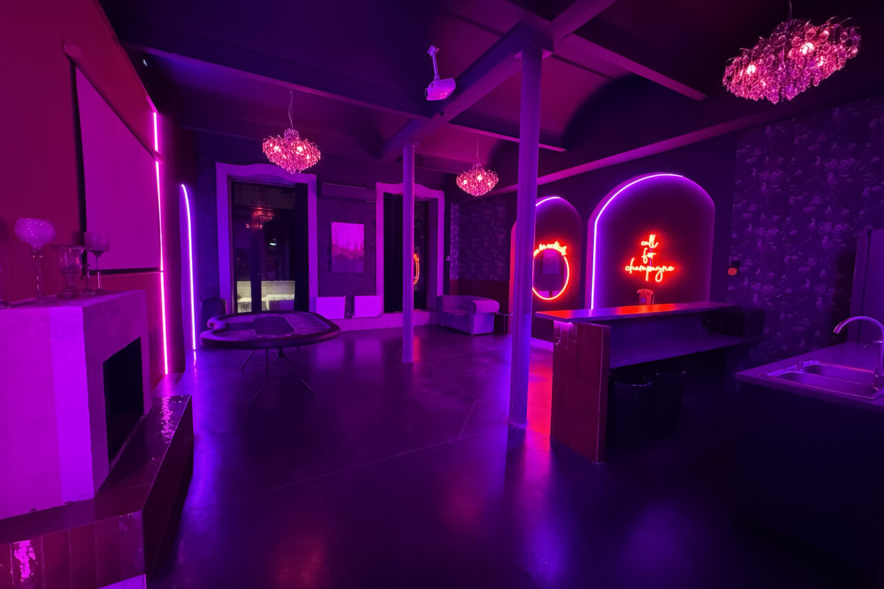Арендовать Белый лофт с яркой мебелью. Лофт-пространство с открытой планировкой, стол, барная стойка, раковина, камин. Неоновые вывески на стене, полумрак, фиолетовая и розовая подсветка, подвесные светильники, оттенки тёмных цветов, зеркала, большие окна.