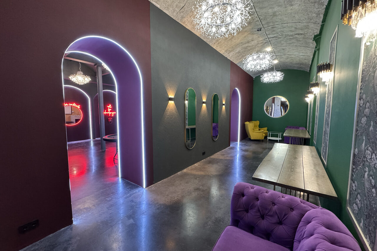 Арендовать Белый лофт с яркой мебелью. Просторное помещение с высокими арочными проходами, фиолетовым диваном и желтым креслом. На стенах неоновые вывески и круглое зеркало. Освещение из подвесных светильников, стены зелёного и тёмно-серого цвета. Пол тёмный, гладкий.