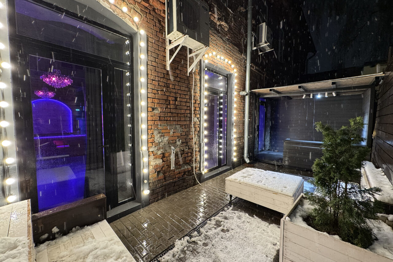 Арендовать Белый лофт с яркой мебелью. Уличное пространство с кирпичными стенами, освещённое гирляндами. Две двери, одна с фиолетовой подсветкой внутри. На полу снег. Белые деревянные скамейки и ящики с растениями. Сверху навес, кондиционеры на стене.