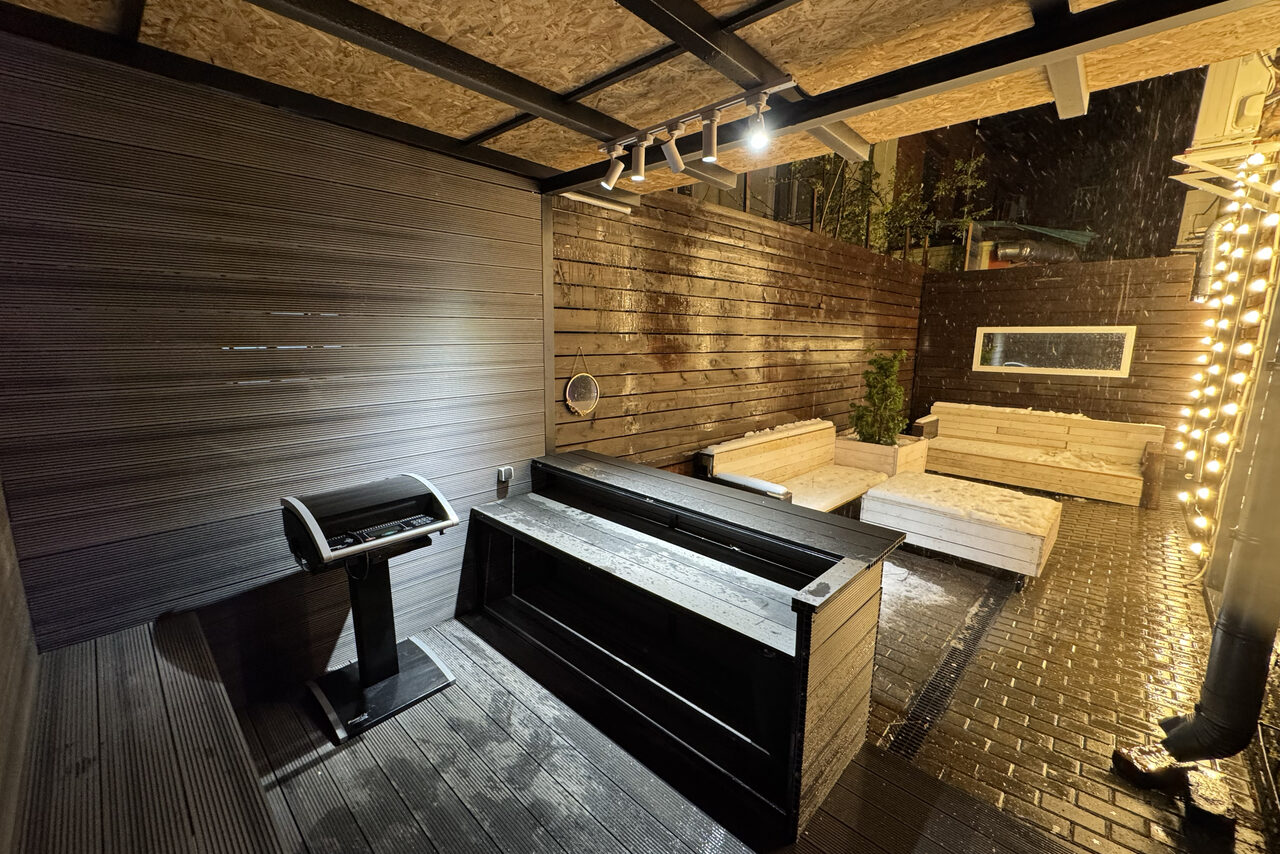 Арендовать Белый лофт с яркой мебелью. Уличное пространство с деревянными стенами и полом, две светлые скамьи, тёмный стол и стойка. Висит световая гирлянда на правой стороне, в центре потолка — прожектор. На улице темно, светло-коричневые стены.